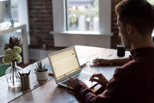 Man sitting at a desk using his laptop, from over his side shoulder.